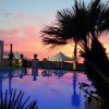 Piscina al tramonto con palme e gazebo, cielo rosa e arancione riflesso nell'acqua.