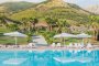 Piscina tranquilla con ombrelloni bianchi, circondata da palme e colline verdi.