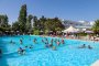 Piscina affollata con adulti e bambini, circondata da alberi e strutture.