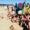 Bambini giocano in spiaggia con una mascotte verde.