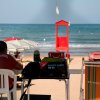 Uomo seduto di fronte al mare, con ombrelloni e torretta rossa.