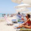 Spiaggia affollata con ombrelloni bianchi e sdraio, donna in costume sorseggia una bevanda rossa.