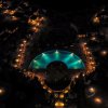 Vista notturna dall'alto di un resort illuminato con piscina centrale e vialetti circondati da luci.