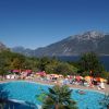 Piscina con persone e ombrelloni colorati, circondata da montagne e lago.