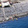 Spiaggia affollata con ombrelloni e lettini, vista dall'alto. Canoe e pedalò sulla riva.