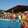 Una piscina affollata con persone sul bordo. Un tetto in legno nelle vicinanze e case sullo sfondo.