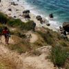 Sentiero scende verso una spiaggia rocciosa con persone e ombrelloni colorati.