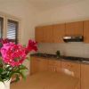 Dettaglio cucina, fiori rosa su tavolo, arredi in legno chiaro, finestra con persiane.