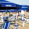 Una spiaggia con sedie a sdraio e ombrelloni blu; persone in background.
