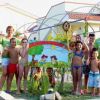 Un gruppo di bambini e adulti sorride davanti a un cartello colorato all'aperto.