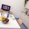 Cucina moderna con tavolo bianco imbandito e un cesto di frutta. Sullo sfondo, una TV accesa.