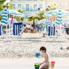 Bambino gioca con secchielli sulla spiaggia, ombrelloni e edificio sullo sfondo.