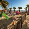 Parco giochi sabbioso con altalene e scivolo, circondato da palme.