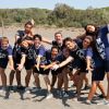 Un gruppo di persone in magliette blu posano sorridenti sulla spiaggia con le braccia aperte.