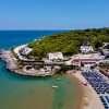Una spiaggia sabbiosa con ombrelloni blu e un promontorio alberato vicino al mare.