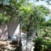 Parte di una casa nascosta da alberi verdi, con una porta e ombre di foglie sul terreno.