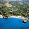 Una spiaggia curva con sabbia chiara, colline verdi e mare blu intenso.