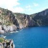 Scogliera rocciosa che si affaccia su un mare blu, con vegetazione sulla cima e cielo sereno.