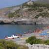 Spiaggia affollata con ombrelloni colorati, bagnanti in acqua e scogliere sullo sfondo.