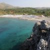Baia con acqua cristallina, vegetazione rigogliosa sulla riva e rocce in primo piano.
