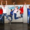 Persone che saltano insieme sul palco, indossando magliette colorate e jeans in una sala interna.