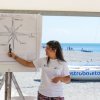 Persona insegna la rosa dei venti su un cartellone in spiaggia, con una barca sullo sfondo.
