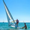 Una persona fa windsurf mentre qualcuno la aiuta in acqua. Una barca a vela sullo sfondo.