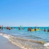 Spiaggia affollata con bagnanti e vele in mare sotto un cielo azzurro.