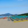 Spiaggia con lettini, ombrelloni turchesi, pedalò rossi. Montagne sullo sfondo e mare calmo.