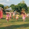 Bambini vestiti da fate corrono in un parco giochi con uno scivolo rosso, circondati da alberi.