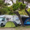 Camper e tenda posizionati sotto alberi ombreggiati.