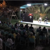 Un pubblico seduto all'aperto guarda due persone su un palco illuminato di sera.