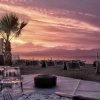Un tramonto in spiaggia con tavolo apparecchiato, bicchieri e palma. Ombrelloni chiusi sullo sfondo.