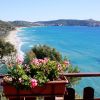 Un balcone con fiori rosa affacciato su una spiaggia con mare blu e colline in lontananza.