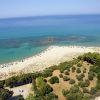 Una spiaggia di sabbia bianca con ombrelloni, mare turchese, alberi e un parcheggio vicino.
