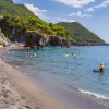 Spiaggia con persone che nuotano e montagne verdi sullo sfondo.