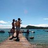 Persone su un molo di legno guardano un kite sul lago, con barche sullo sfondo.