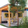 Bungalow con terrazza coperta, sedie bianche, albero e cespuglio in primo piano.