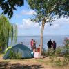 Una tenda da campeggio vicino a un lago, persone in piedi e un cielo soleggiato.
