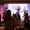 Persone danzano su un palco illuminate da luci, con un pubblico.