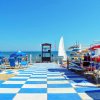 Spiaggia con ombrelloni blu, pedana a scacchi bianchi e blu, e canoe colorate vicino al mare.