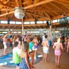 Persone ballano in un locale con tetto in legno. Presente una palla da discoteca e un bancone bar.
