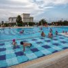 Piscina all'aperto con persone che nuotano e bagnino a bordo vasca.