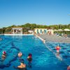 Piscina affollata con persone che nuotano e prendono il sole, ombrelloni in fondo.
